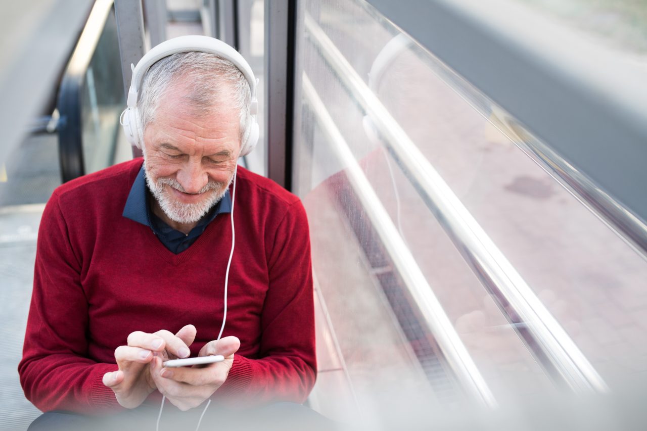 Handsome senior man holding smart phone, headphones on his ears, listening music, sitting in glass passage.