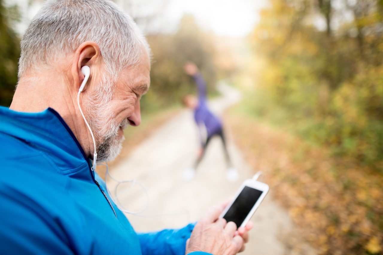 Senior runners in nature, stretching. Man with smart phone with earphones. Listening music or using a fitness app. Using phone app for tracking weight loss progress, running goal or summary of his run.