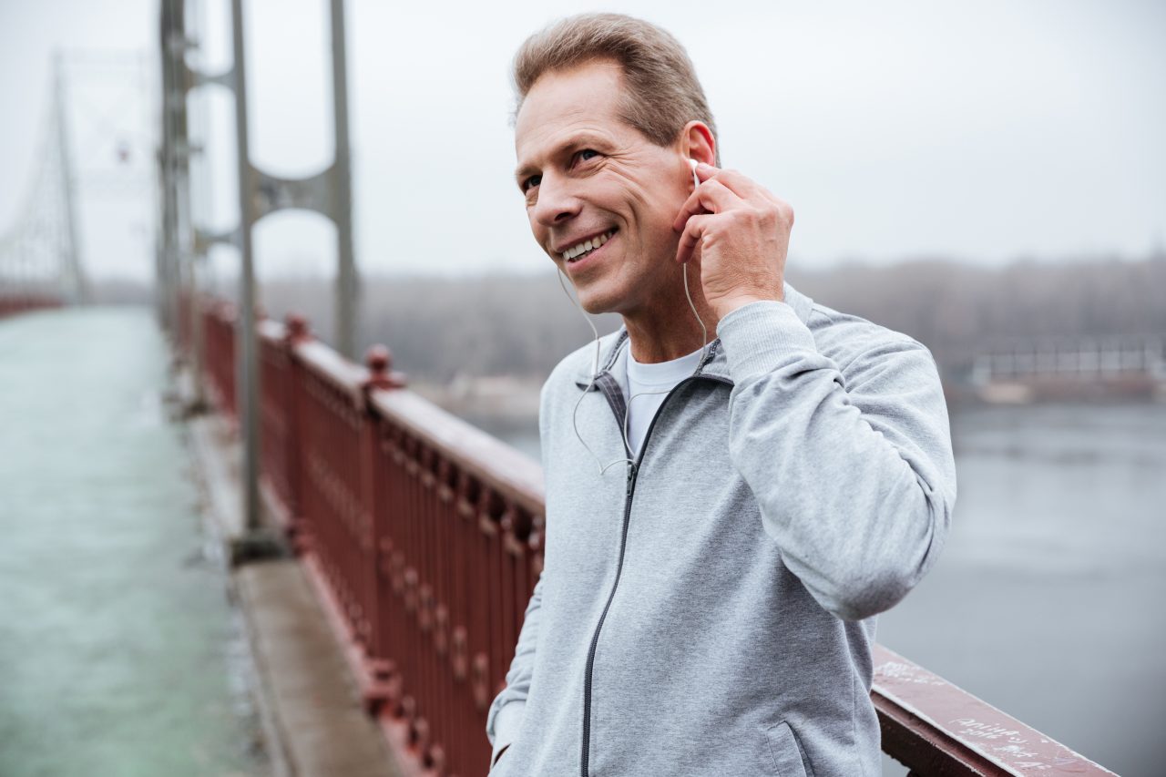 Smiling Runner in gray sportswear standing and listening to music on bridge