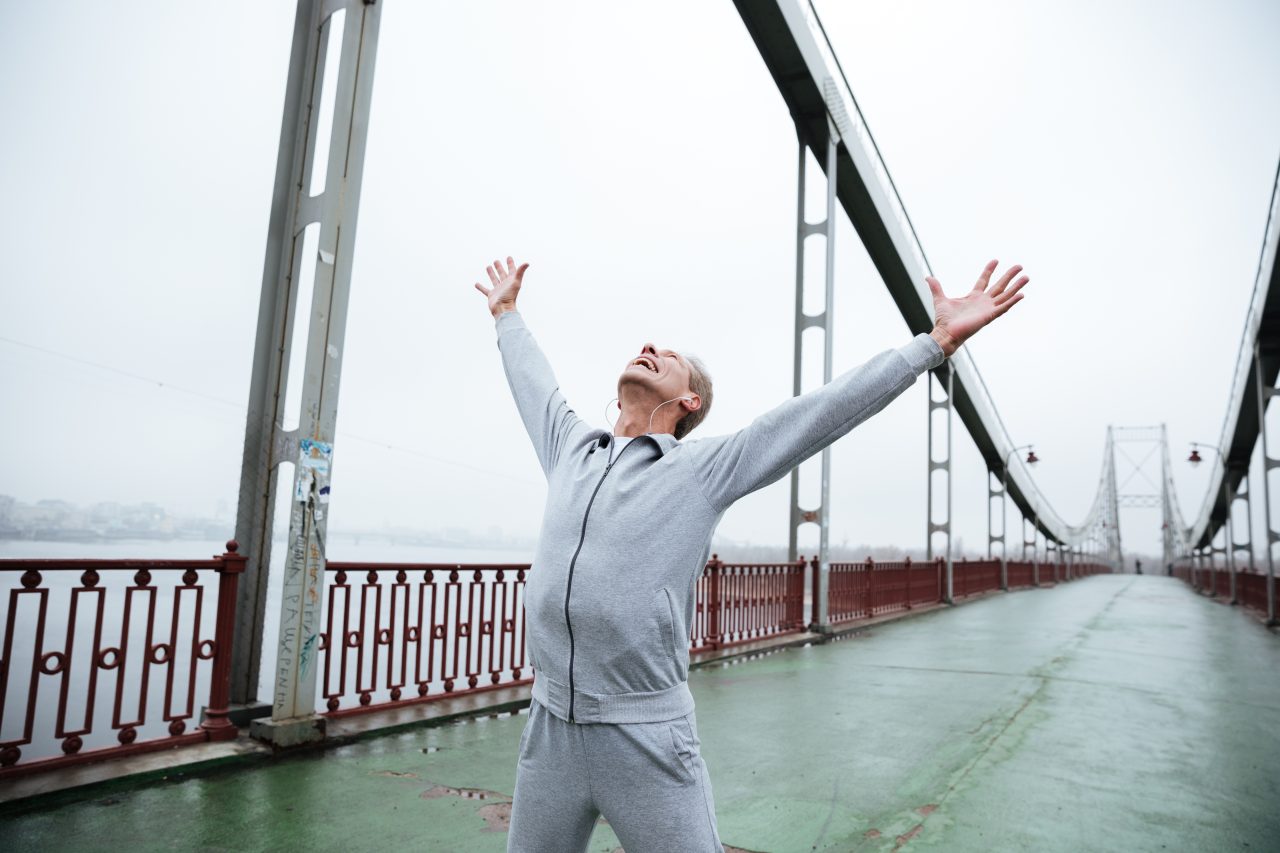 Happy elderly man in gray sportswear standing on bridge with hands up looking up
