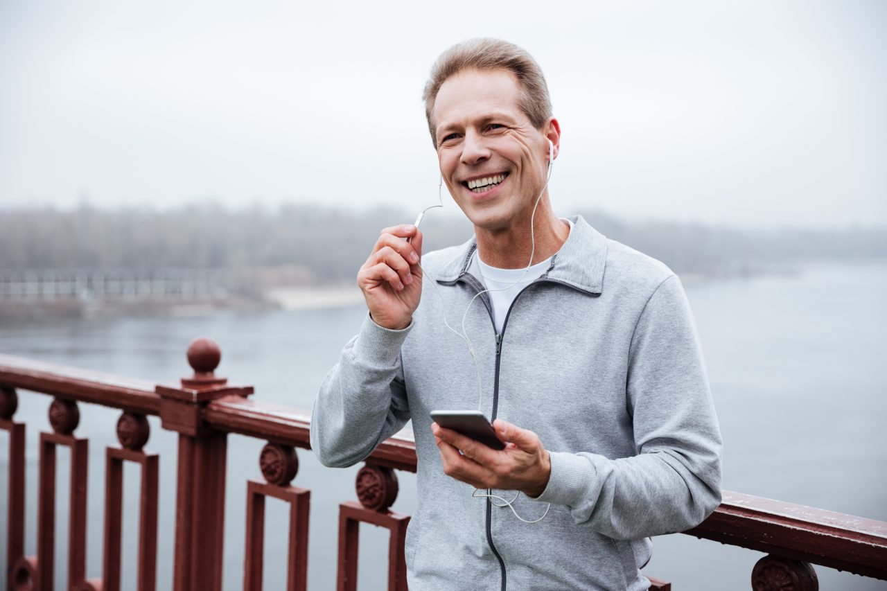 Smiling Runner in gray sportswear holding phone and listening to music on bridge