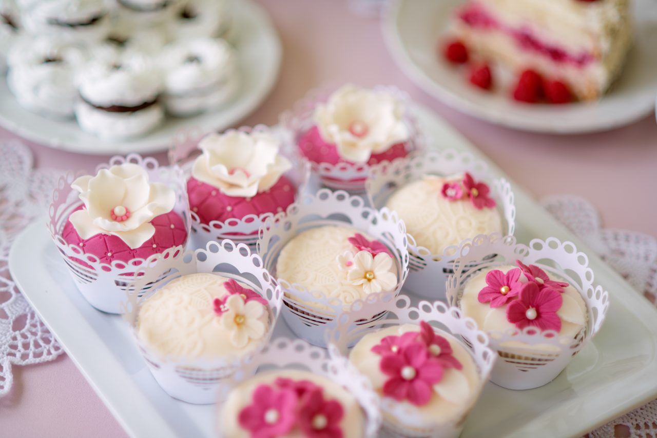 Close up, cupcakes on tray decorated with pink flowers laid on table with pink tablecloth and handmade lace. Candy bar.